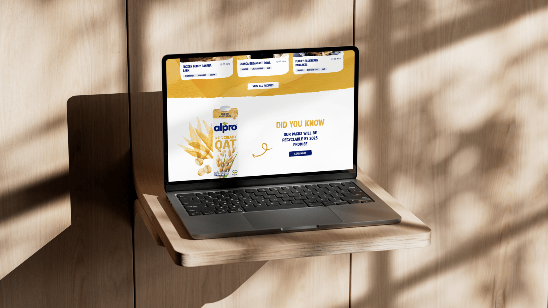 A laptop displaying an Alpro oat milk product webpage sits on a wooden floating shelf with dramatic shadow patterns cast on the wall.