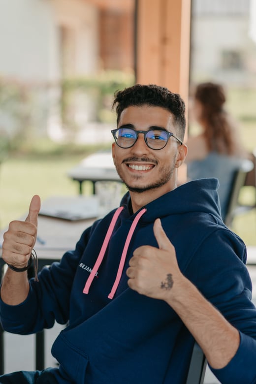 A teammate wearing glasses gives two enthusiastic thumbs up outdoors.