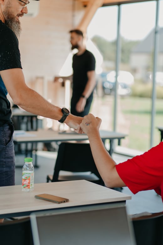 Two teammates shake hands across a desk in a bright office.